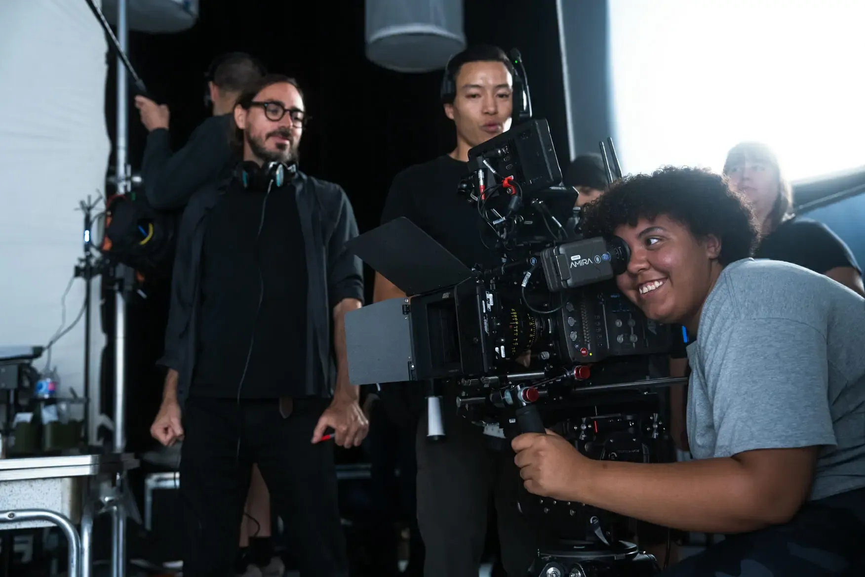 A camera operator smiles into the eyepiece of a cinema camera while the crew looks on