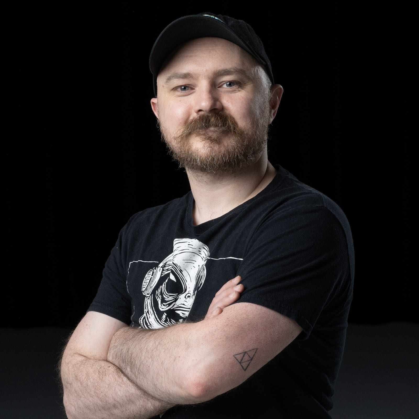 Portrait of Jesse Kellestine, arms folded, in a cap and a black T-shirt
