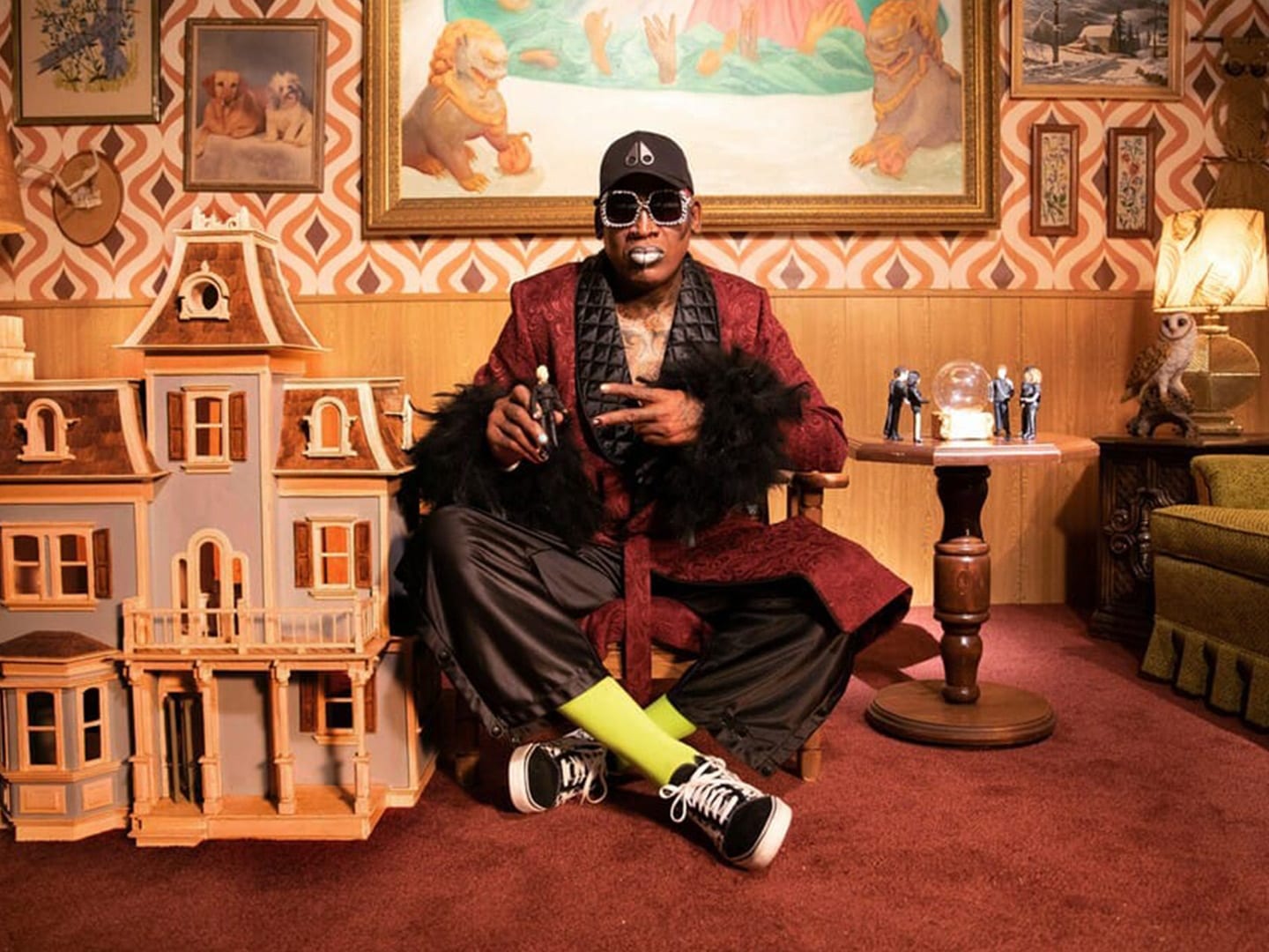 A performer in a burgundy robe seated on the floor of a wood panelled set beside a doll house