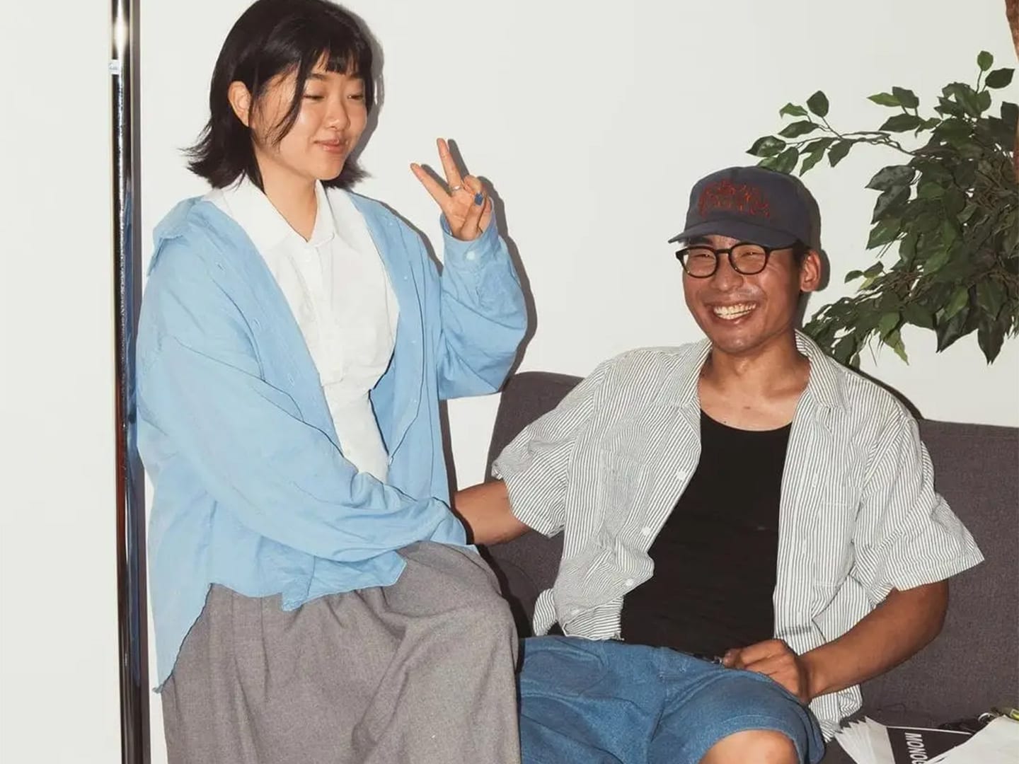 Two people laughing on a sofa beside a plant, one throwing a peace sign