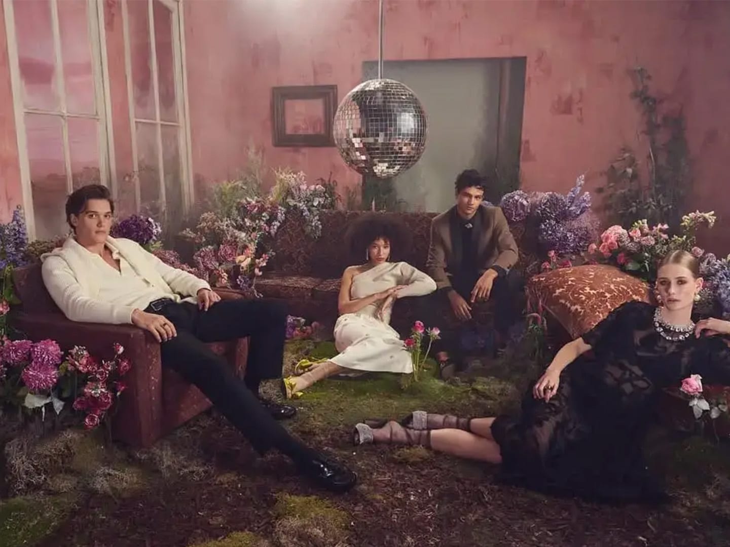 A group draped across sofas and moss under a mirror ball on a pink lit set