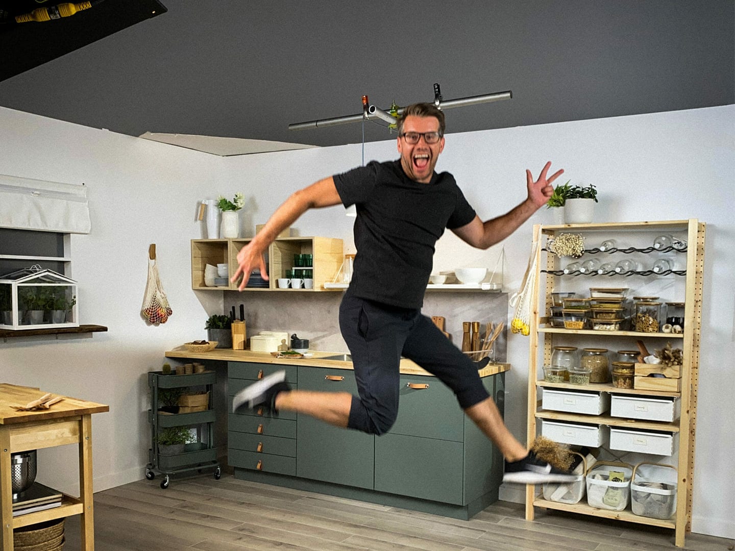 A presenter caught mid jump in a kitchen set lined with shelves and jars