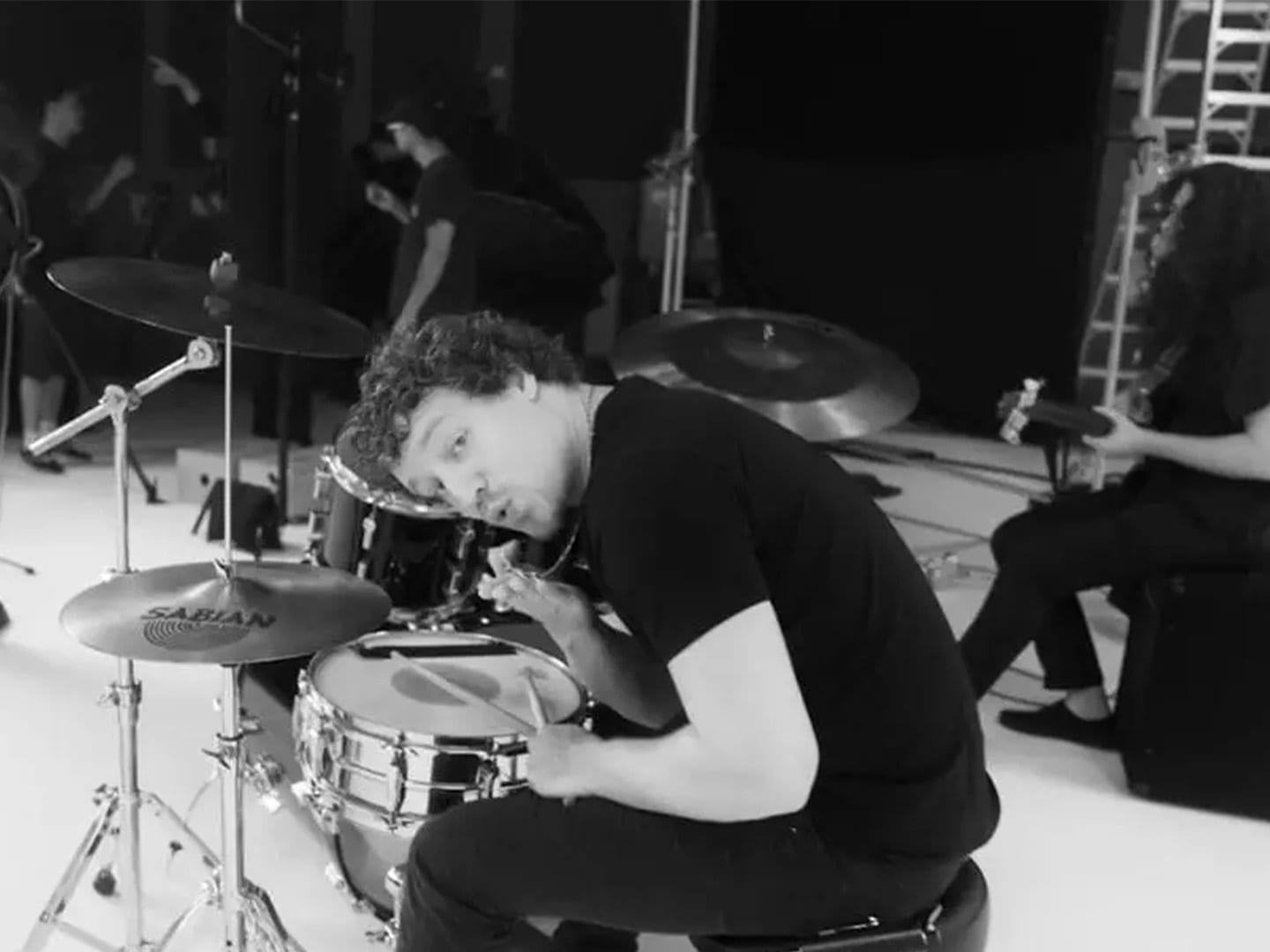 A drummer at the kit in black and white, crew working behind him