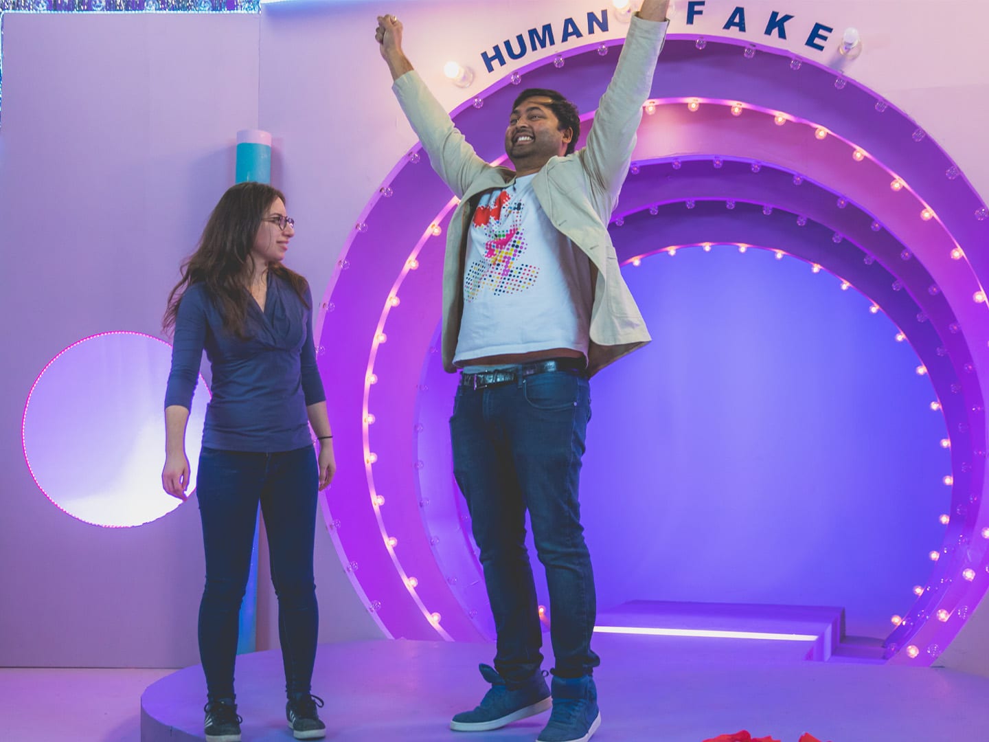 A host with both arms raised in front of a purple neon arch, a crew member beside him
