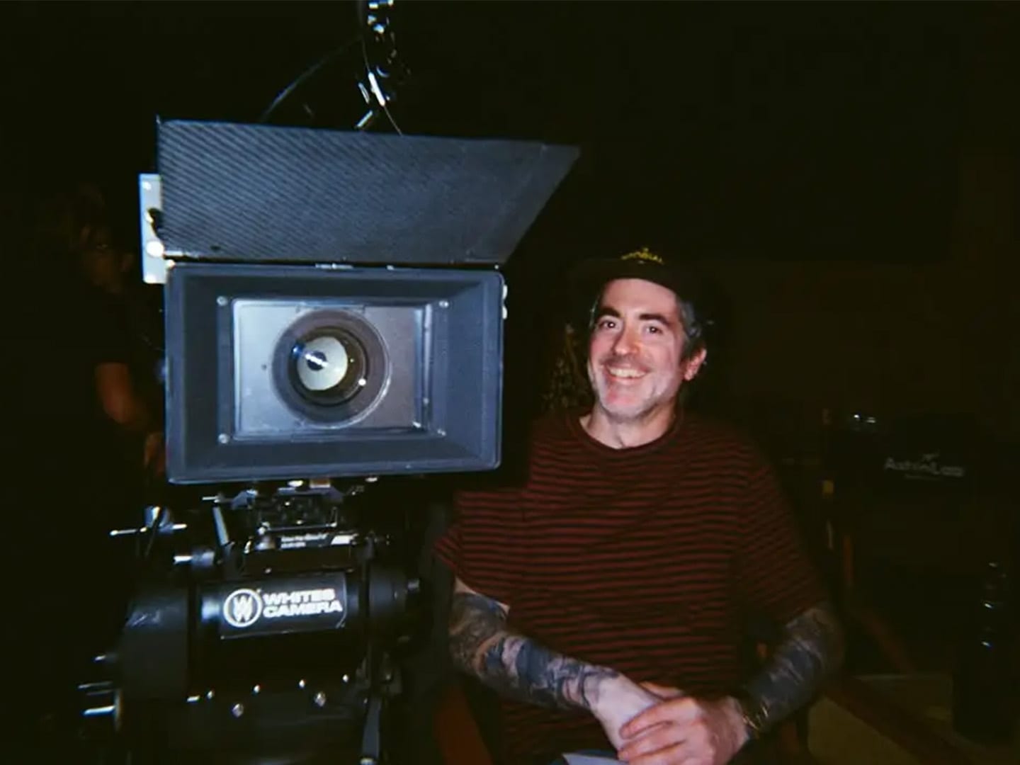 A camera operator grinning behind a cinema camera and matte box in the dark