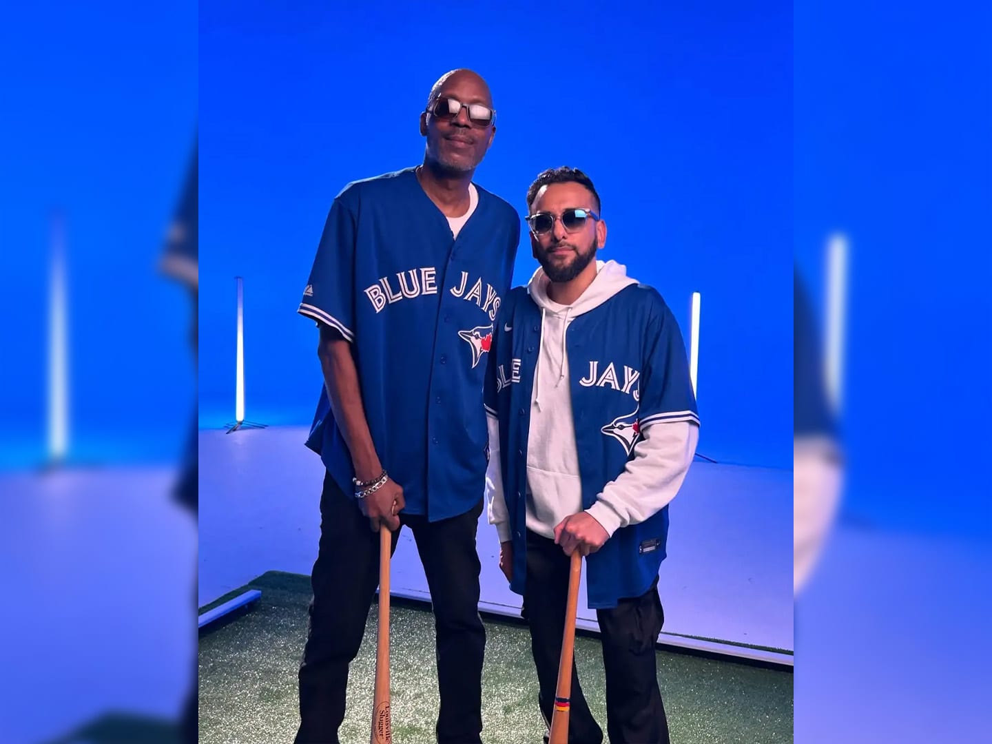 Two performers in Blue Jays gear, one holding a bat, on a blue lit set