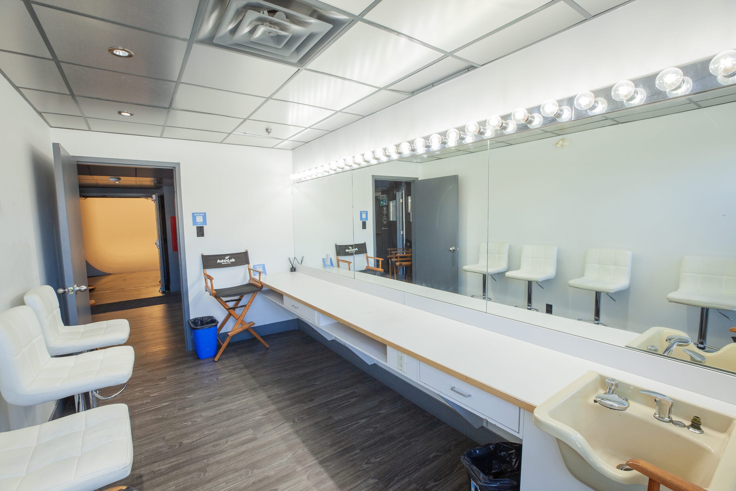 The hair and makeup room, a long bulb lit mirror over the vanity and a hair sink in the foreground