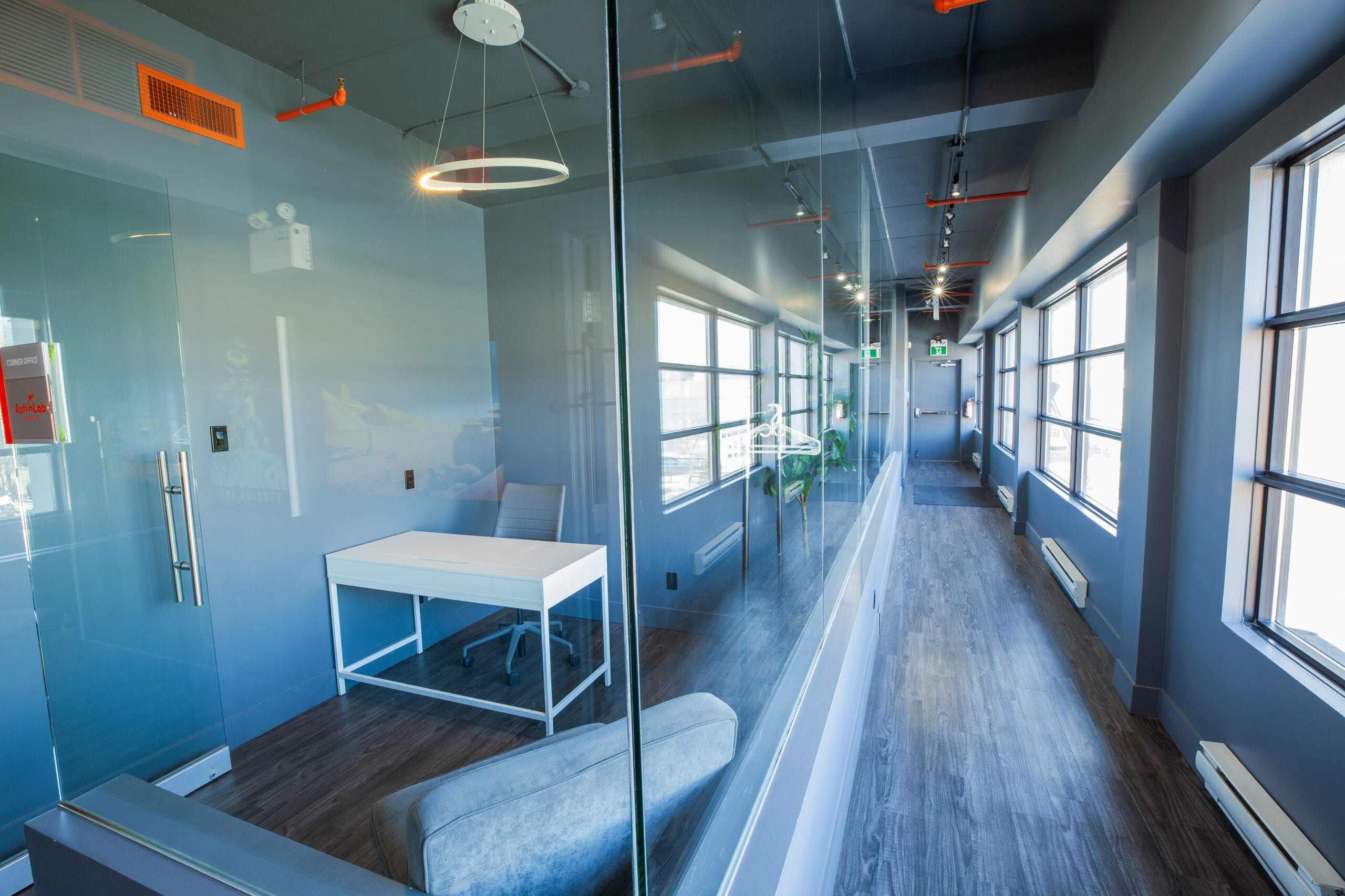 A long, wide hallway with a wall of windows on one side and a glass-walled office on the other